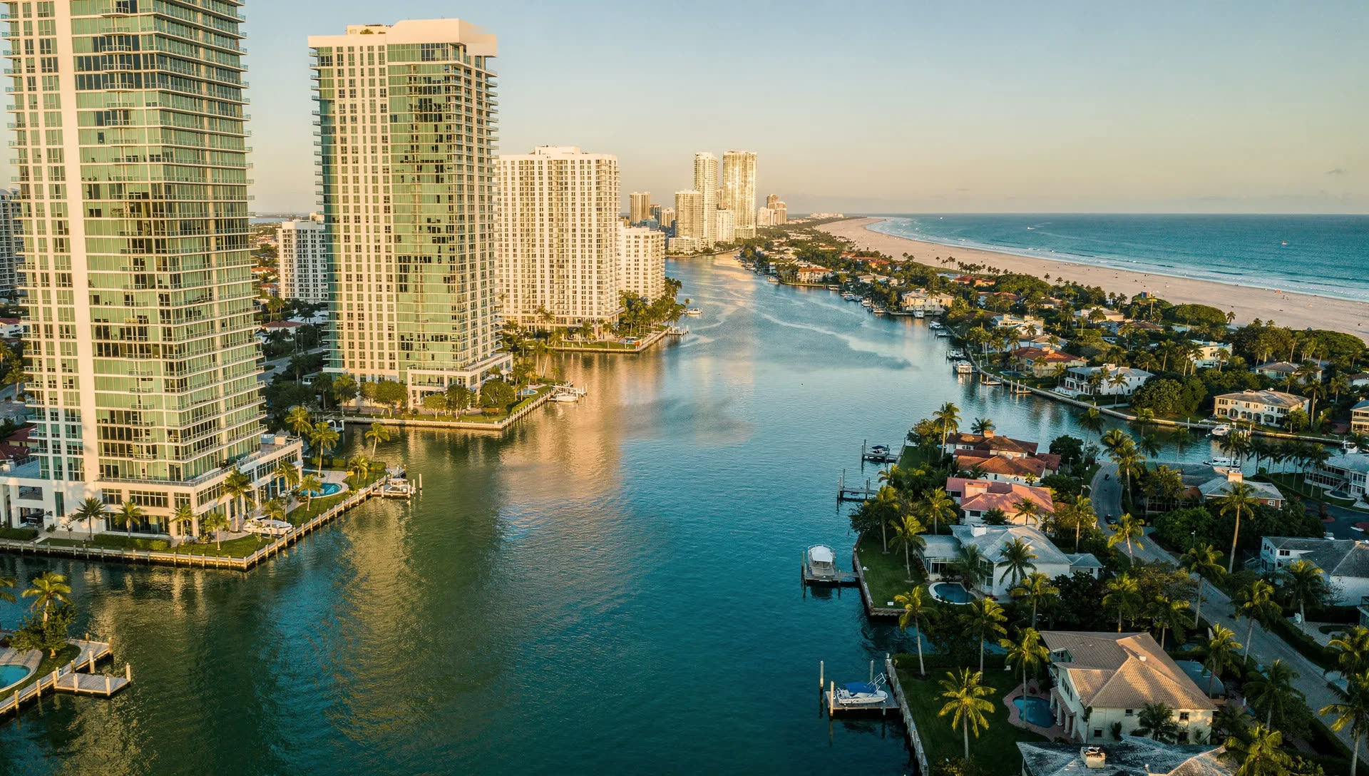 Aerial view at golden hour of high-rise towers along the Intracoastal Waterway, canal-front homes with private docks on the right, and the Atlantic beach on the barrier island beyond.